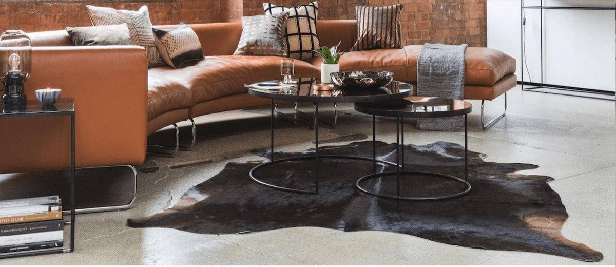 A modern living room scene featuring a large black and brown cowhide area rug on a concrete floor, styled with a curved brown leather sectional sofa, black metal nesting coffee tables, and decorative pillows.